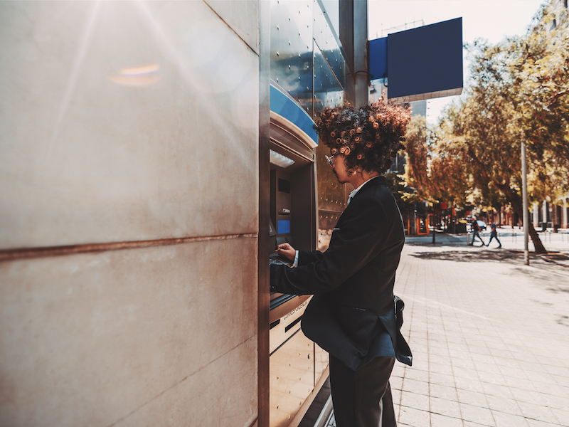A side view of a curly fancy businessman in a formal suit using an outdoor cash dispenser to withdraw money from his account, with a copy space place on the left for your text or logo (A side view of a curly fancy businessman in a formal suit using an