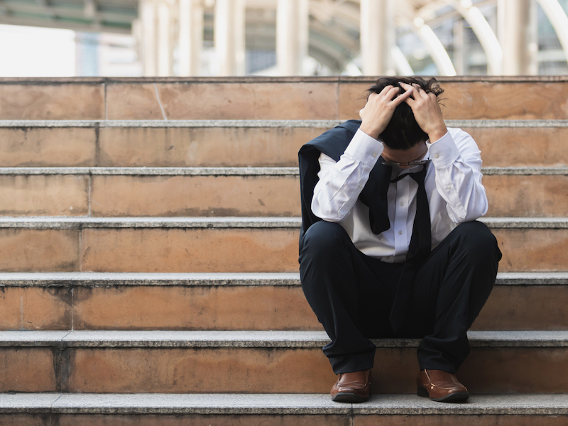 Upset stressed young Asian business man in suit with hands on head sitting on stairs. Unemployment and layoff concept.