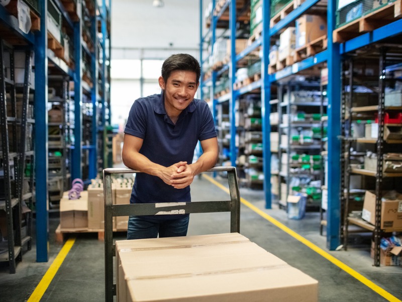 Male worker standing with a trolley with boxes in warehouse. Warehouse worker in uniform with a cart in aisle of storage racks.