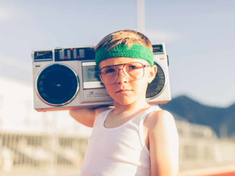 A young boy dressed in headband and retro workout attire is having a great time listening to music on his boombox.