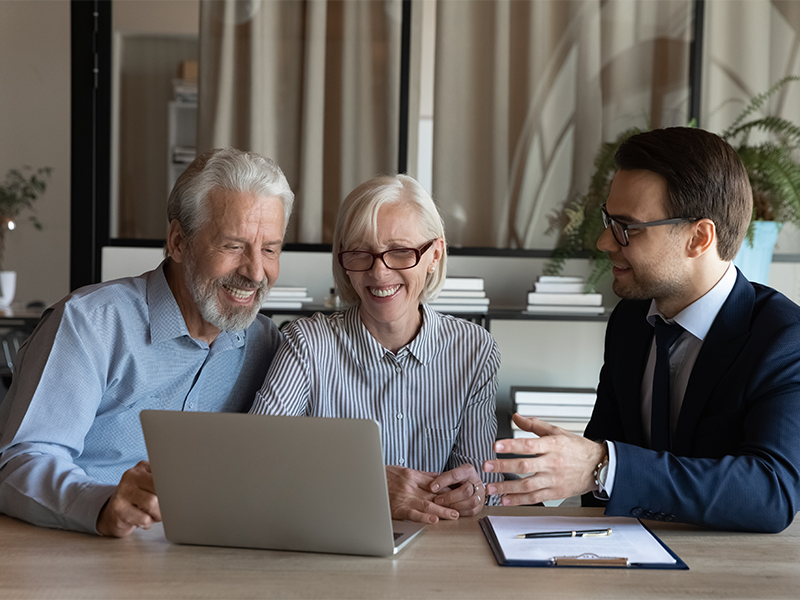Smiling senior couple sit with a lawyer around a table looking at a laptop