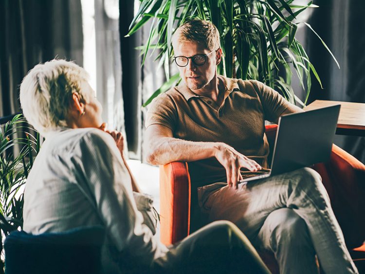 Man sitting cross legged with a laptop on his lap, speaking to a woman