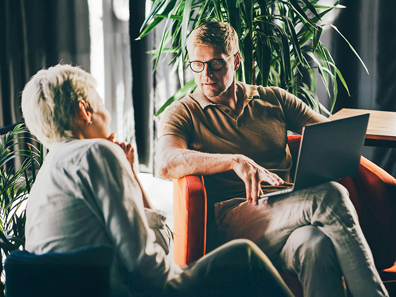 Man sitting cross legged with a laptop on his lap, speaking to a woman