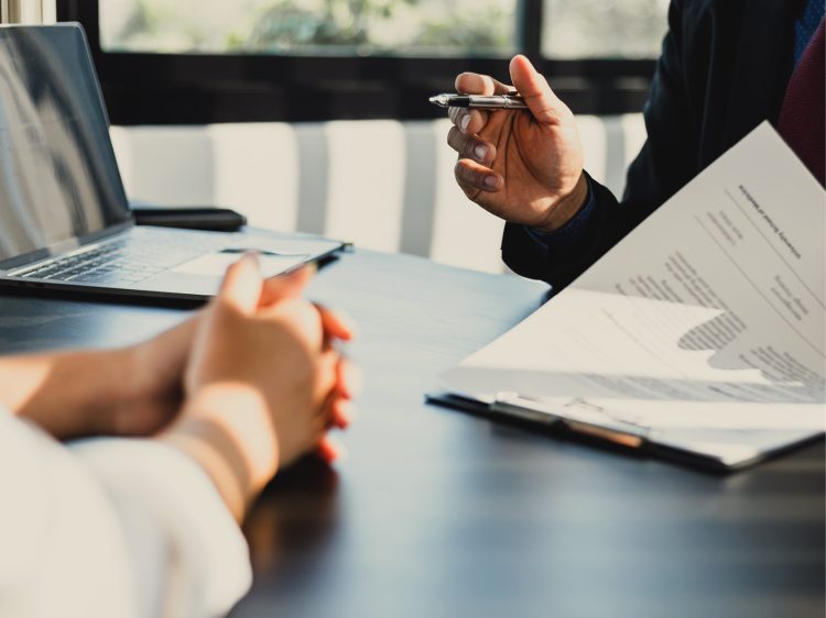 Financial advisor meeting with a client at a modern office table. Advisor holds a pen and reviews a printed financial report with charts and graphs, while the client listens with clasped hands. An open laptop is on the table, sunlight streaming through large windows.