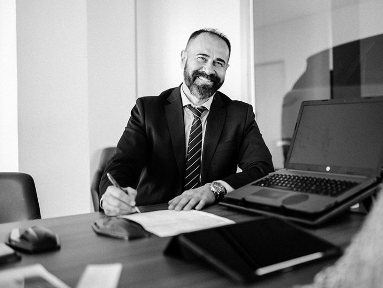 Man in suit and tie, sitting at desk with papers and laptop, smiling