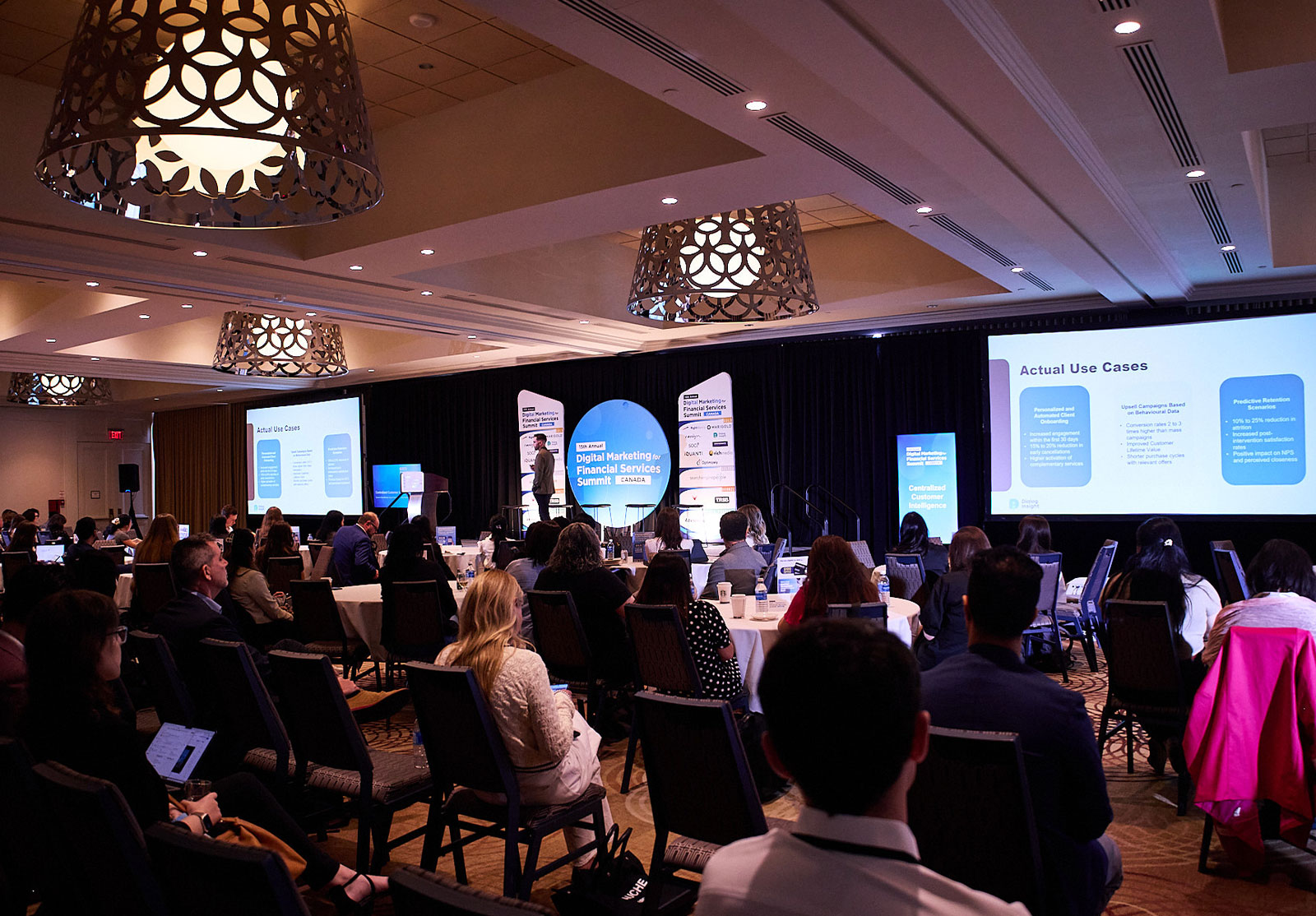 Large conference room with attendees seated at round tables facing a stage, where a speaker presents slides.