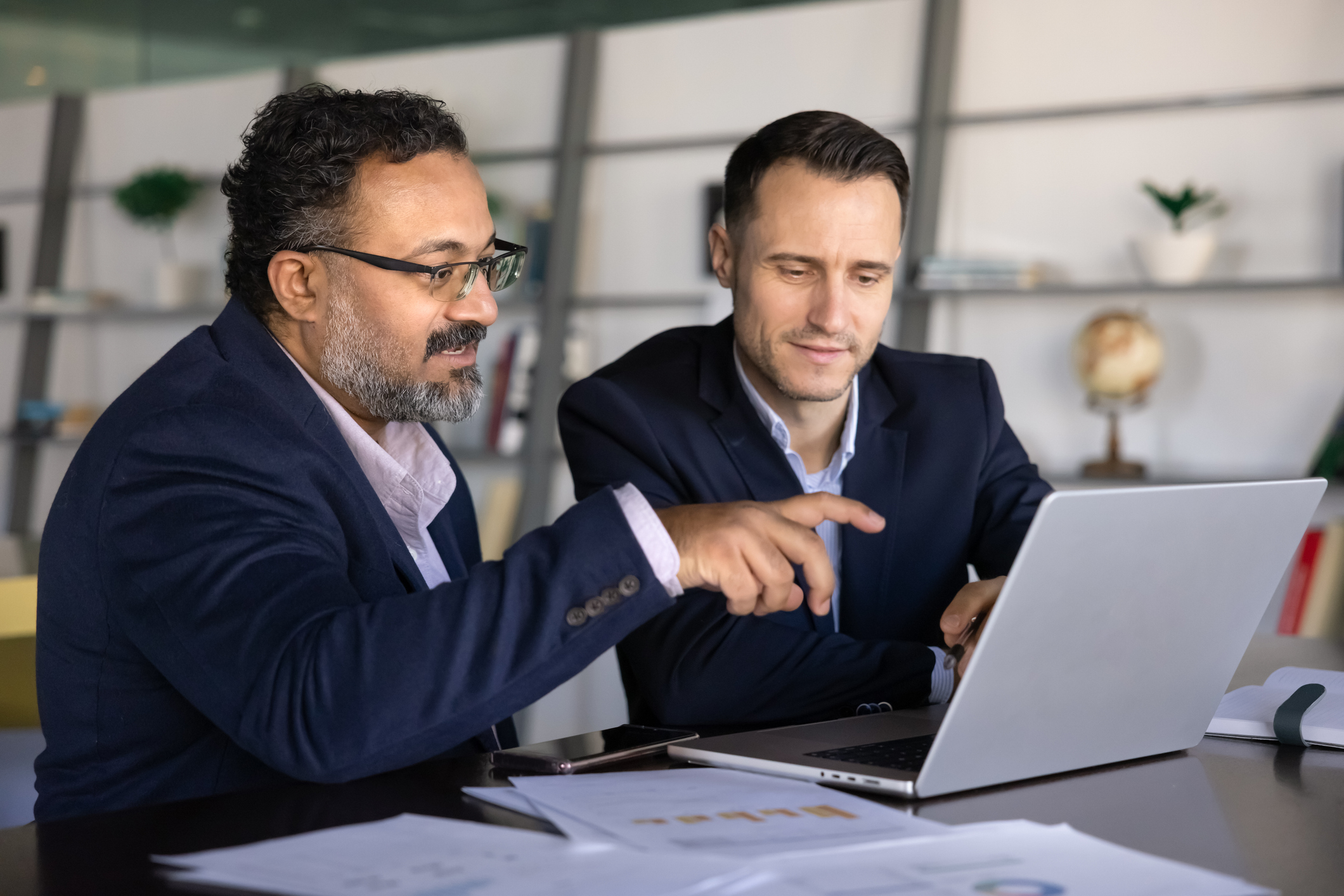 Two diverse advisor businessmen working together at a table, pointing at laptop screen and discussing strategy